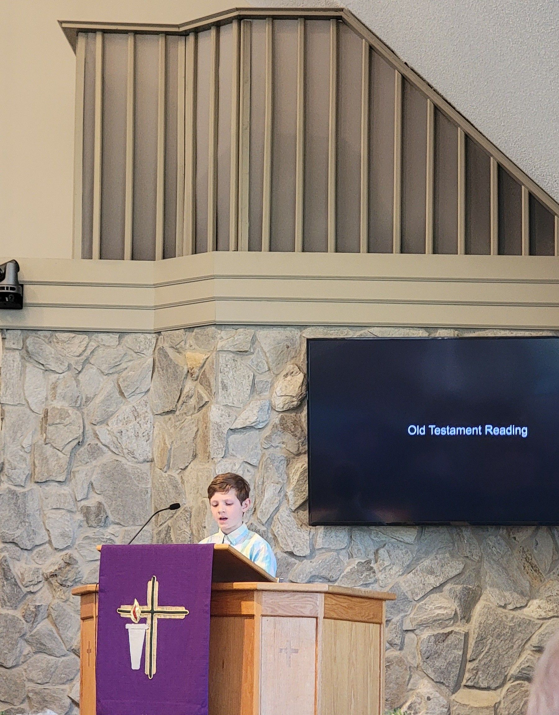 Person speaking at a church lectern with a purple cloth, stone wall and screen behind.