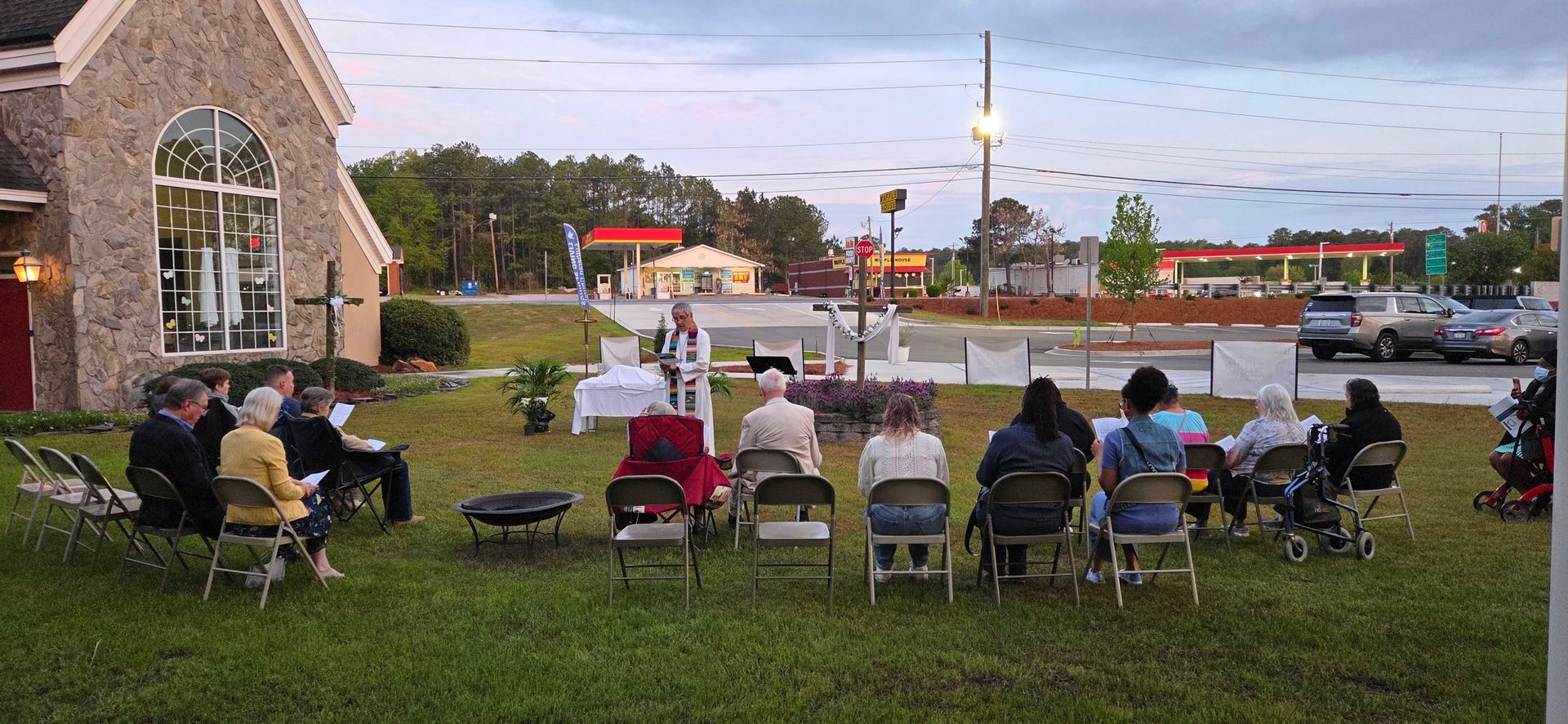 People seated in folding chairs on a lawn watching an outdoor event at dawn.
