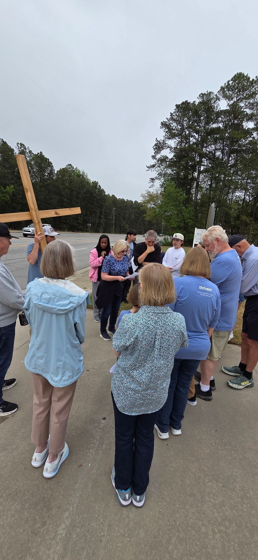 Group gathered outdoors around a raised wooden object, with trees and an overcast sky in the background.