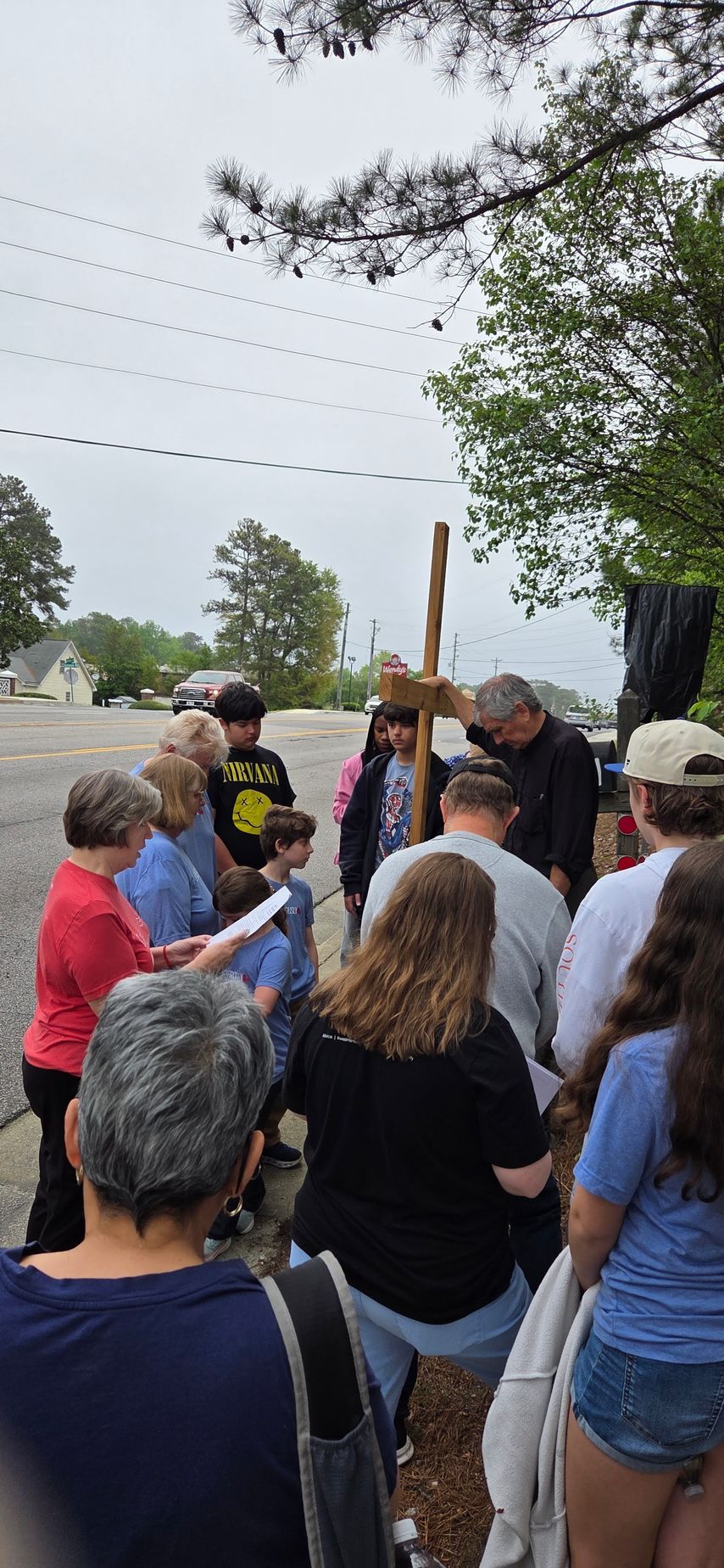Crowd gathered outdoors around a person holding a wooden cross near a roadside