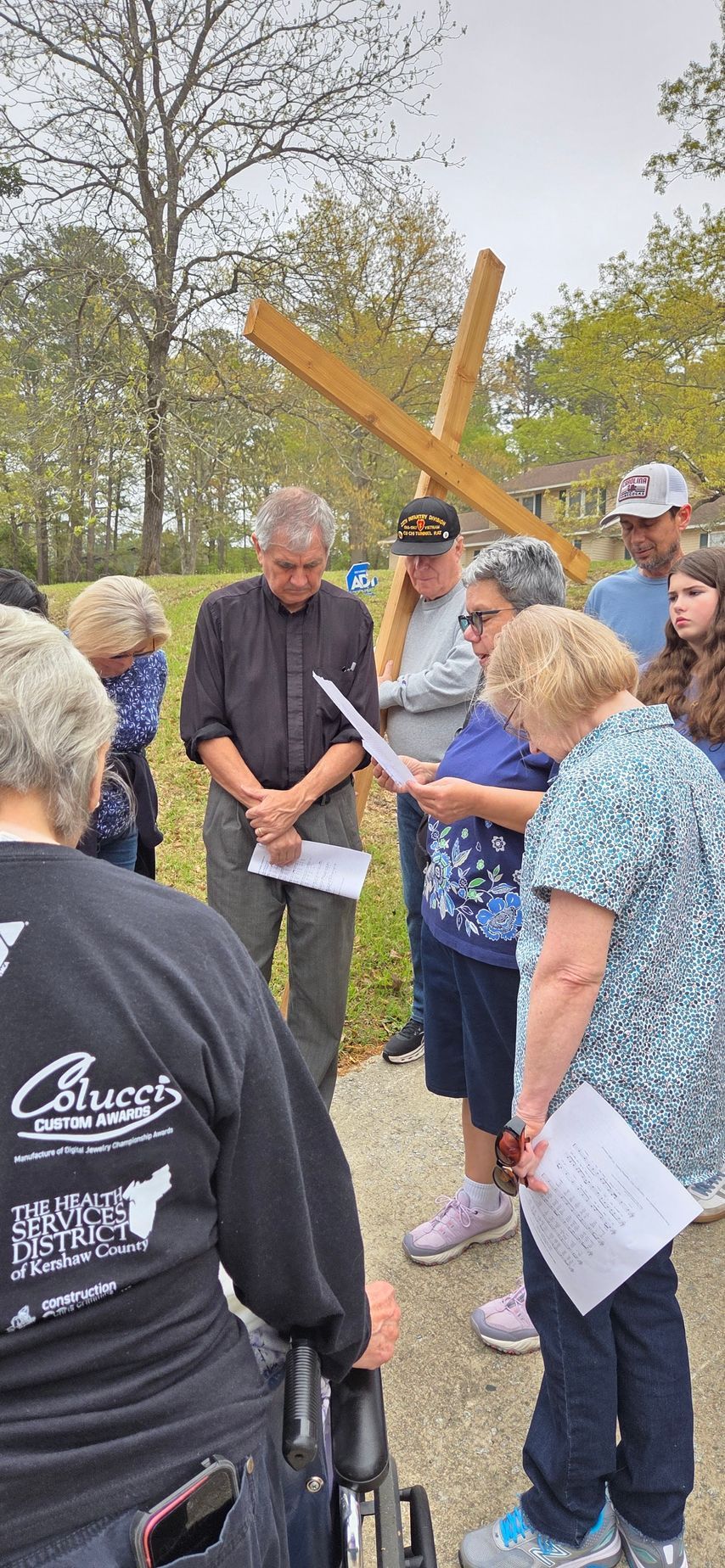 Small group gathered outdoors around a large wooden cross, discussing papers in a park-like setting.