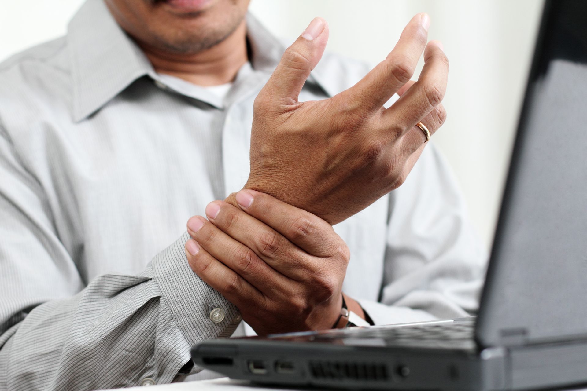 Person in a white shirt holding their wrist beside a laptop.