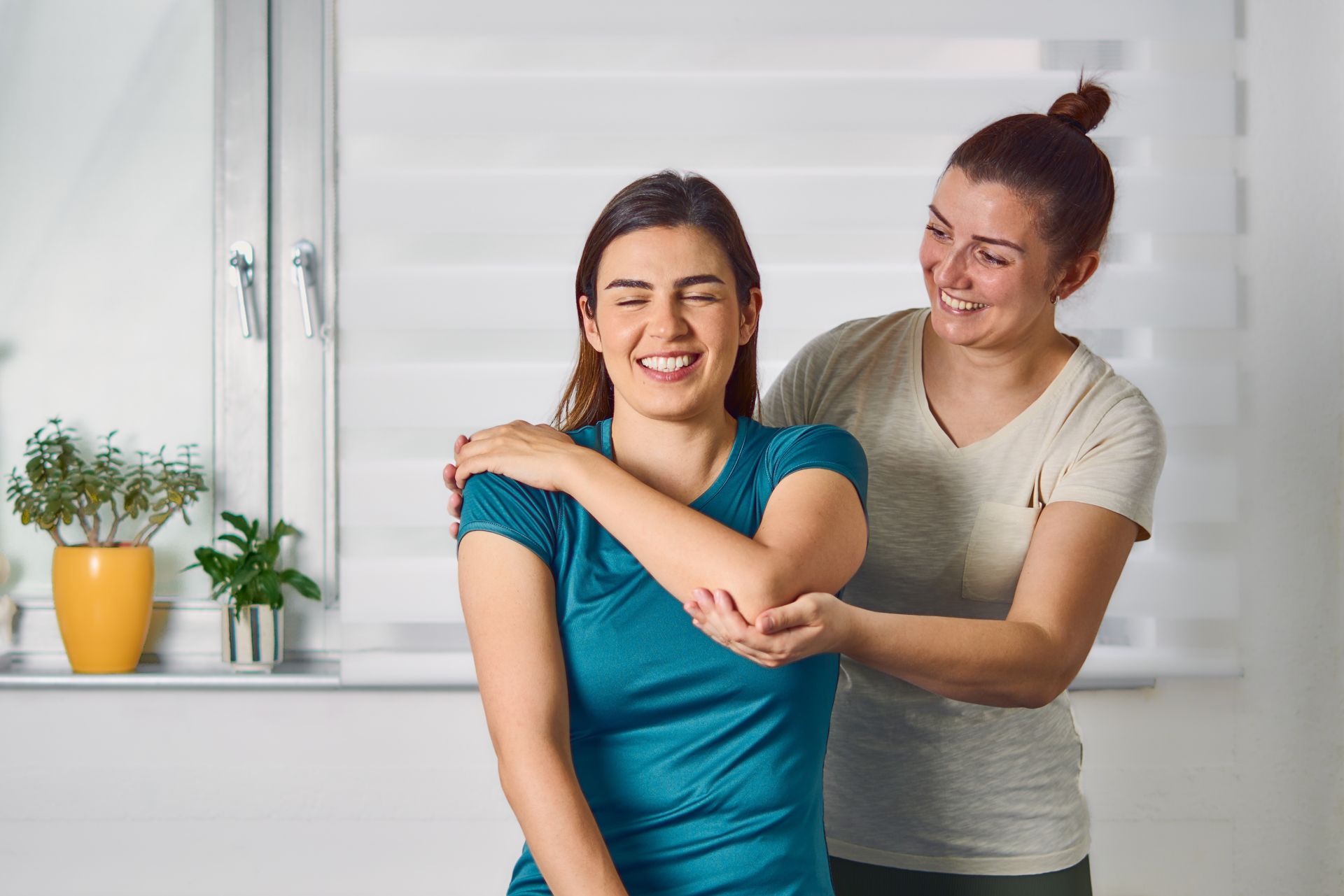 Smiling woman receiving a shoulder massage from another woman in a bright room with plants on the windowsill