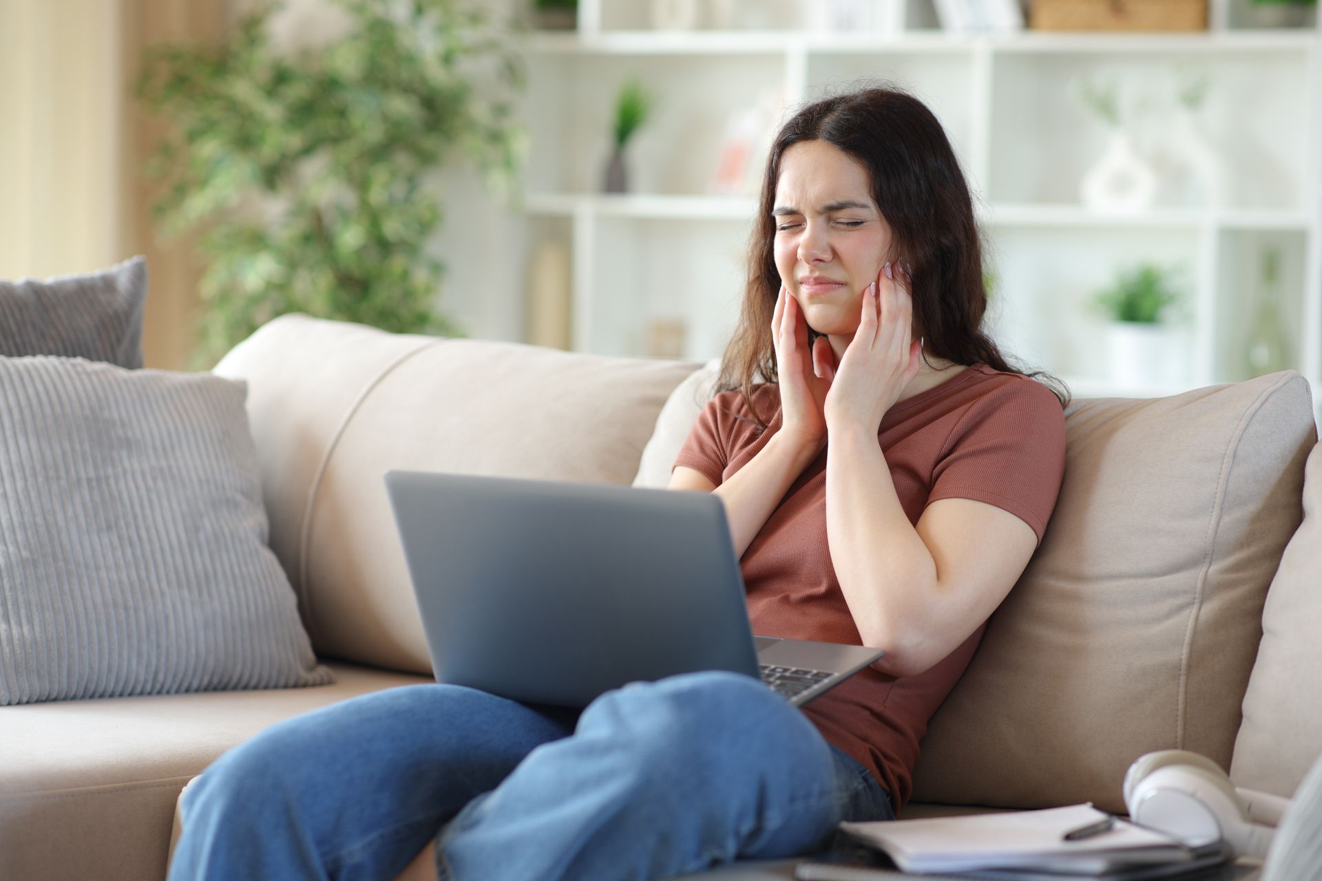 Woman sits on a couch with a laptop, holding her face in discomfort in a bright living room.