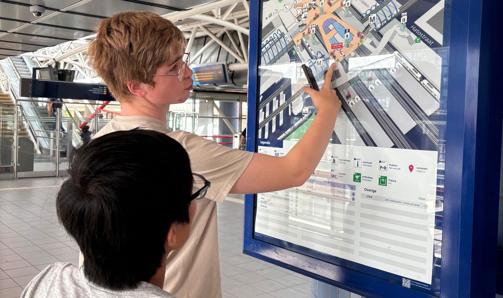 Una persona indica una mappa dei trasporti pubblici su un pannello informativo blu in una stazione.