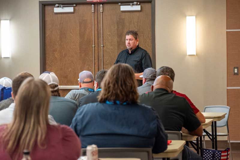 Man speaking to a group in a conference room; attendees seated at tables, listening.
