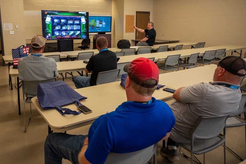 A man leads a presentation in a classroom with screens displaying data. Attendees sit at tables, observing.