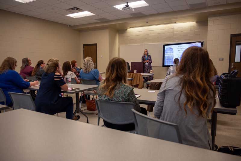 A woman presents at a lecture hall with an audience seated at desks, a screen displaying information.