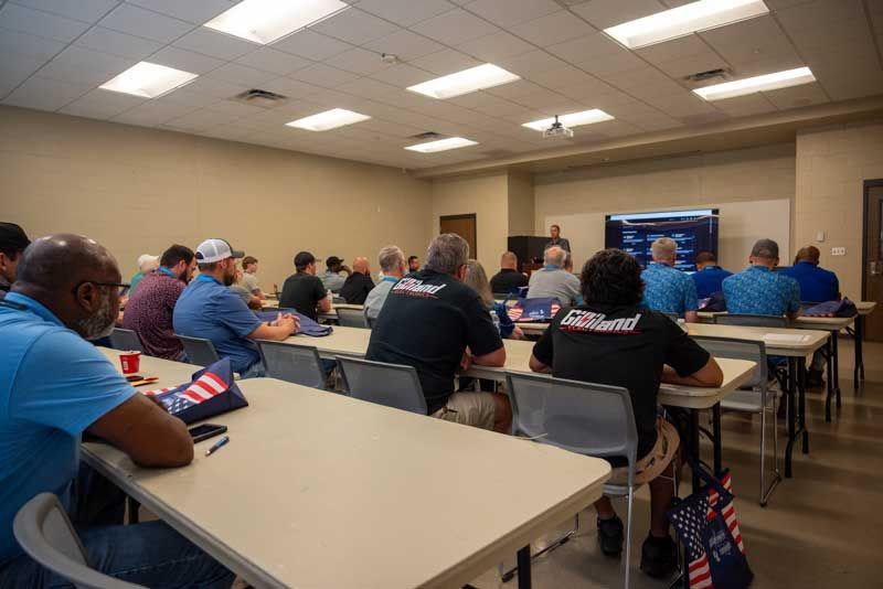 People in a classroom, listening to a presentation on a screen. Some wear blue shirts and black t-shirts.