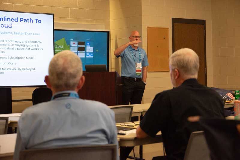 Man giving a presentation, pointing up in a conference room with audience members.
