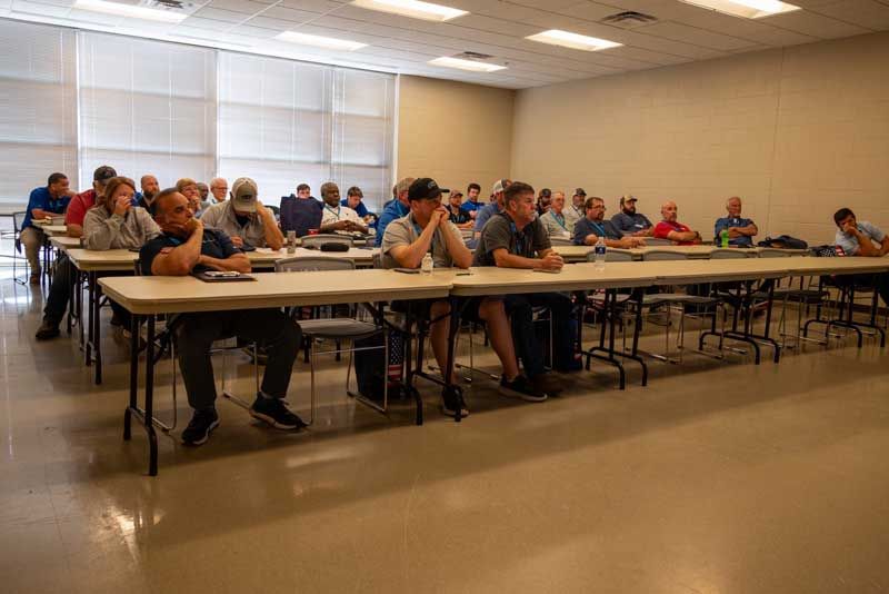 A group of people seated at long tables in a classroom setting, listening attentively.
