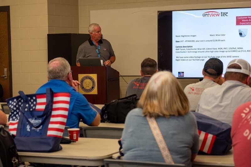 Man presenting at a podium to a seated audience with a presentation screen.