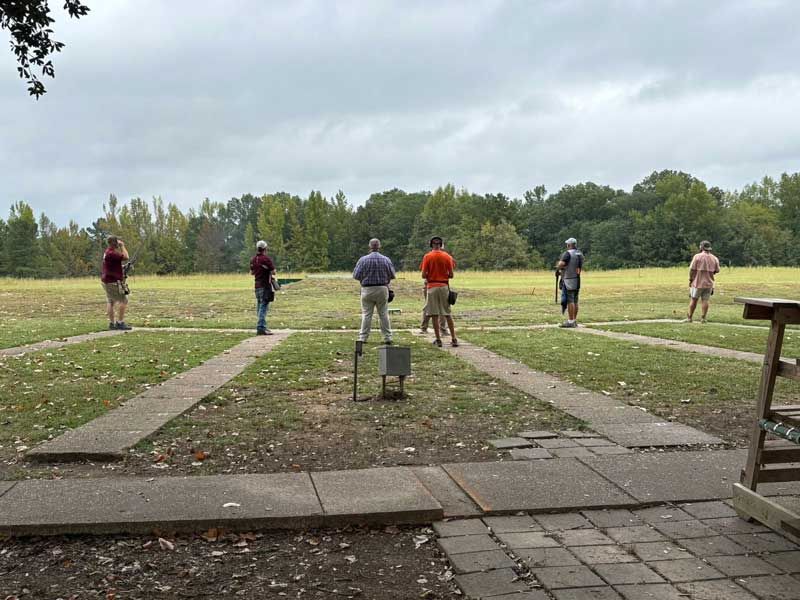 People shooting at a skeet range on a grassy field; overcast sky, trees in the background.