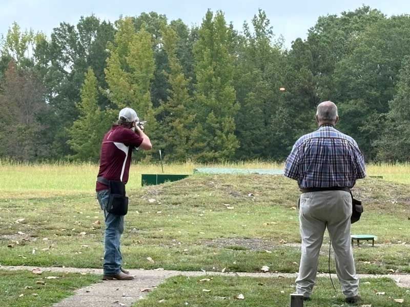 Man shooting shotgun at clay target, another man watches at outdoor range. Green grass, trees.