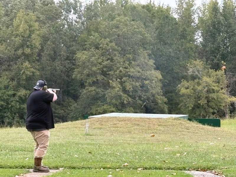 Person shooting shotgun at a clay target on a grassy field.