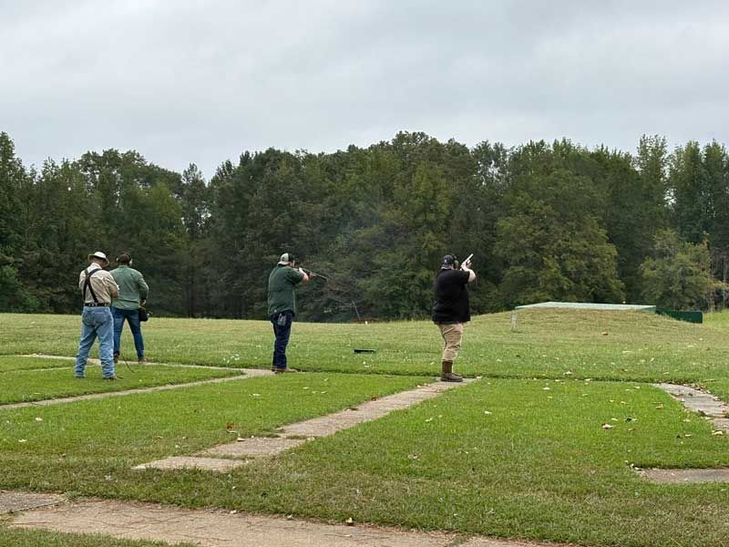 Four people shooting shotguns at an outdoor range, overcast sky, green grass and trees.