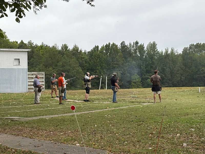 People shooting shotguns at an outdoor range, smoke visible, trees in background.