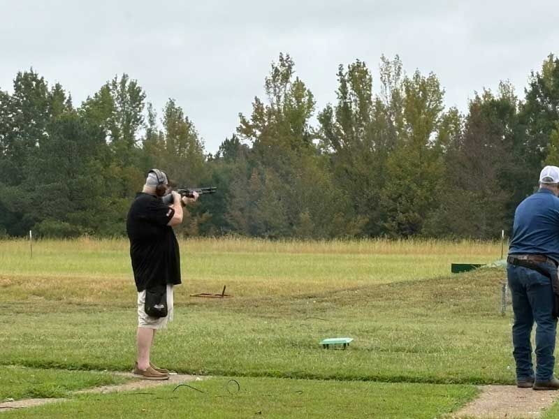 Shooter aims at a target on a grassy field; another man stands nearby. Trees in the background. Overcast sky.