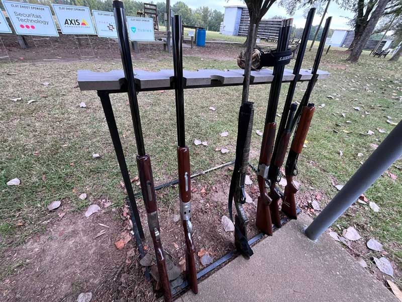 Guns in a rack at a shooting range. Grass, trees, and targets are in the background.
