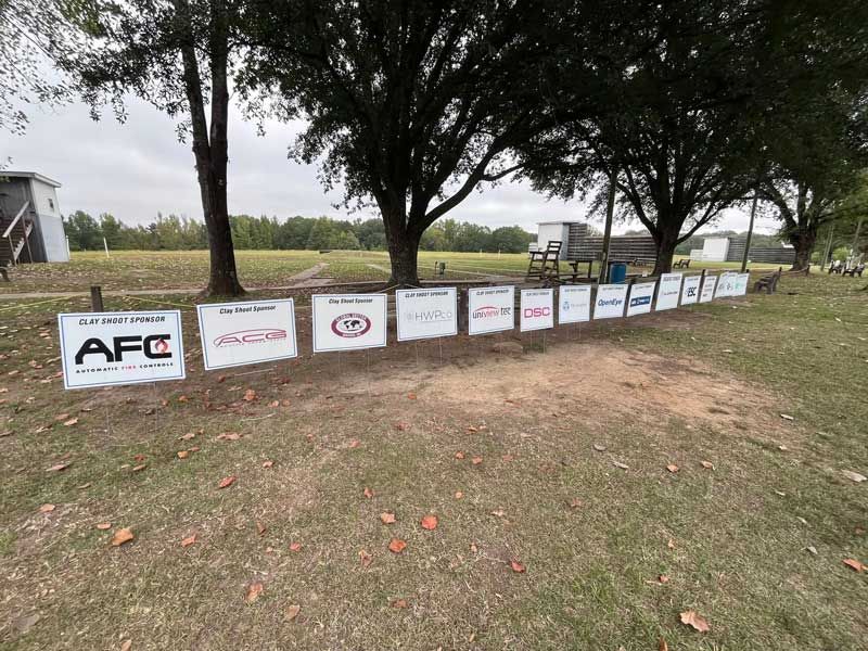 Row of sponsor signs lined up on a grassy field, trees and a shooting range in the background.