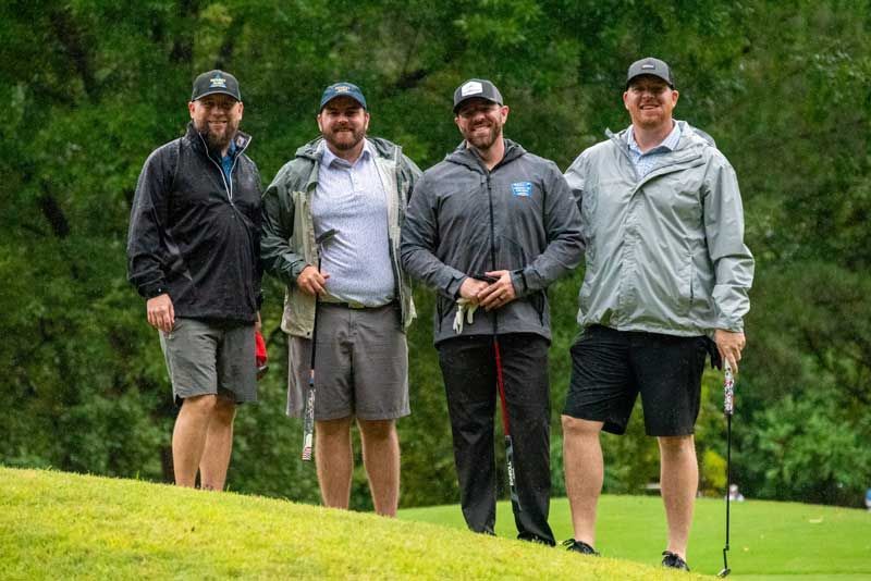 Four men in golf attire pose on a golf course. They wear hats, jackets, and shorts, smiling.