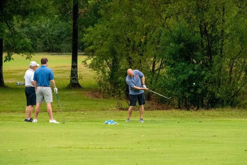 Man swings golf club on green, other men watch in the background.