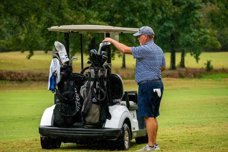 Man at golf cart on green fairway, reaching for a club. Cloudy day, trees in background.