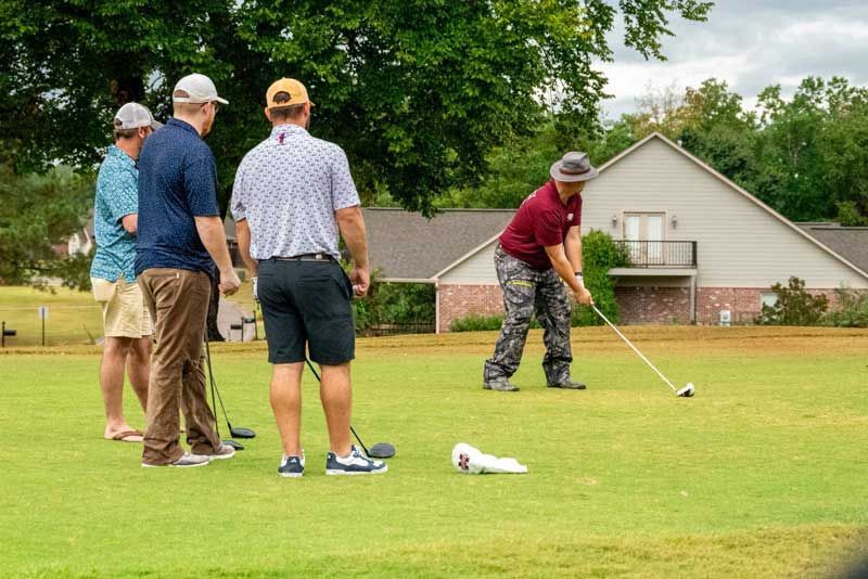 Men playing golf. One tees off as three others watch on a green course. A building is in the background.
