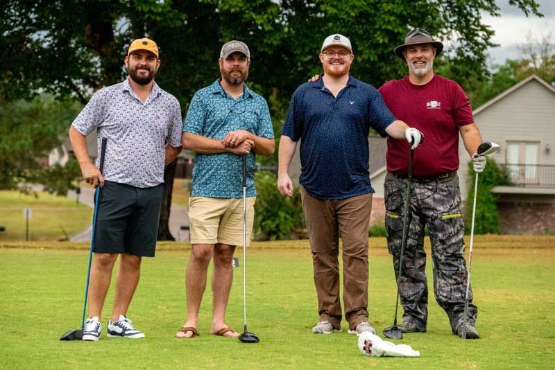 Four men on a golf course pose with clubs. They wear casual clothing, stand on green grass, and smile.