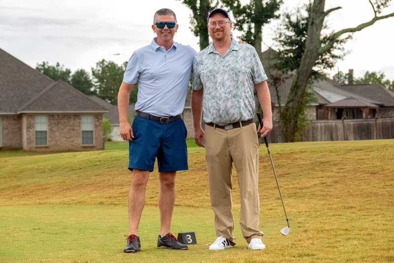 Two men on a golf course. One in blue shorts and the other holding a club. Overcast sky and houses in the background.