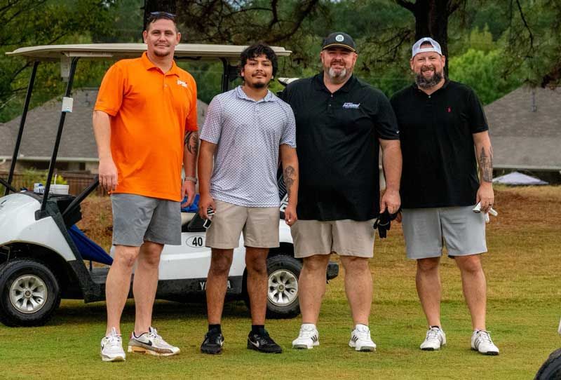 Four men standing on a golf course, near a golf cart. They wear casual clothes in orange, blue, and black.