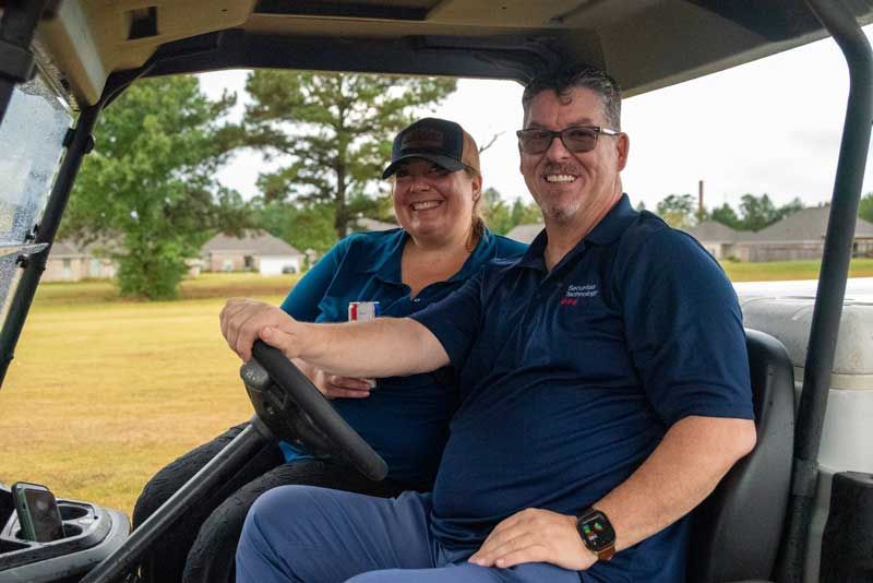 Man smiling while driving golf cart, woman beside him, outdoors.