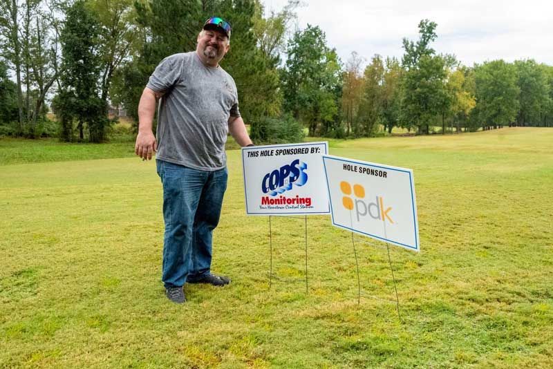 Man standing on a grassy field next to two sponsor signs: MPS Monitoring and pdk. Trees in the background.