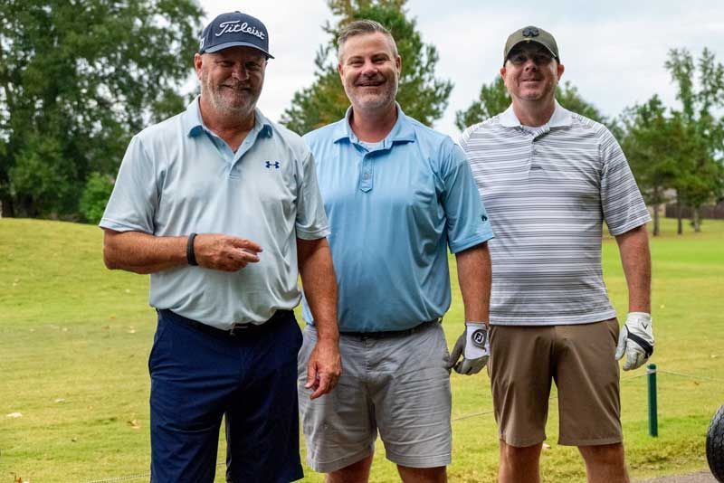 Three men standing on a golf course. They wear golf attire and smile, posing for a photo.
