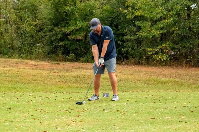 Man playing golf, swinging club on a grassy course with trees in the background.