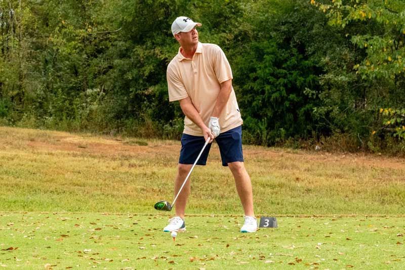 Man in tan shirt and blue shorts swings a golf club on a green course.