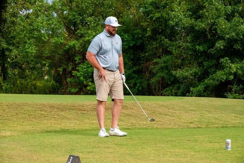 Man in blue shirt and khaki shorts on a golf course, holding a golf club, with trees in the background.