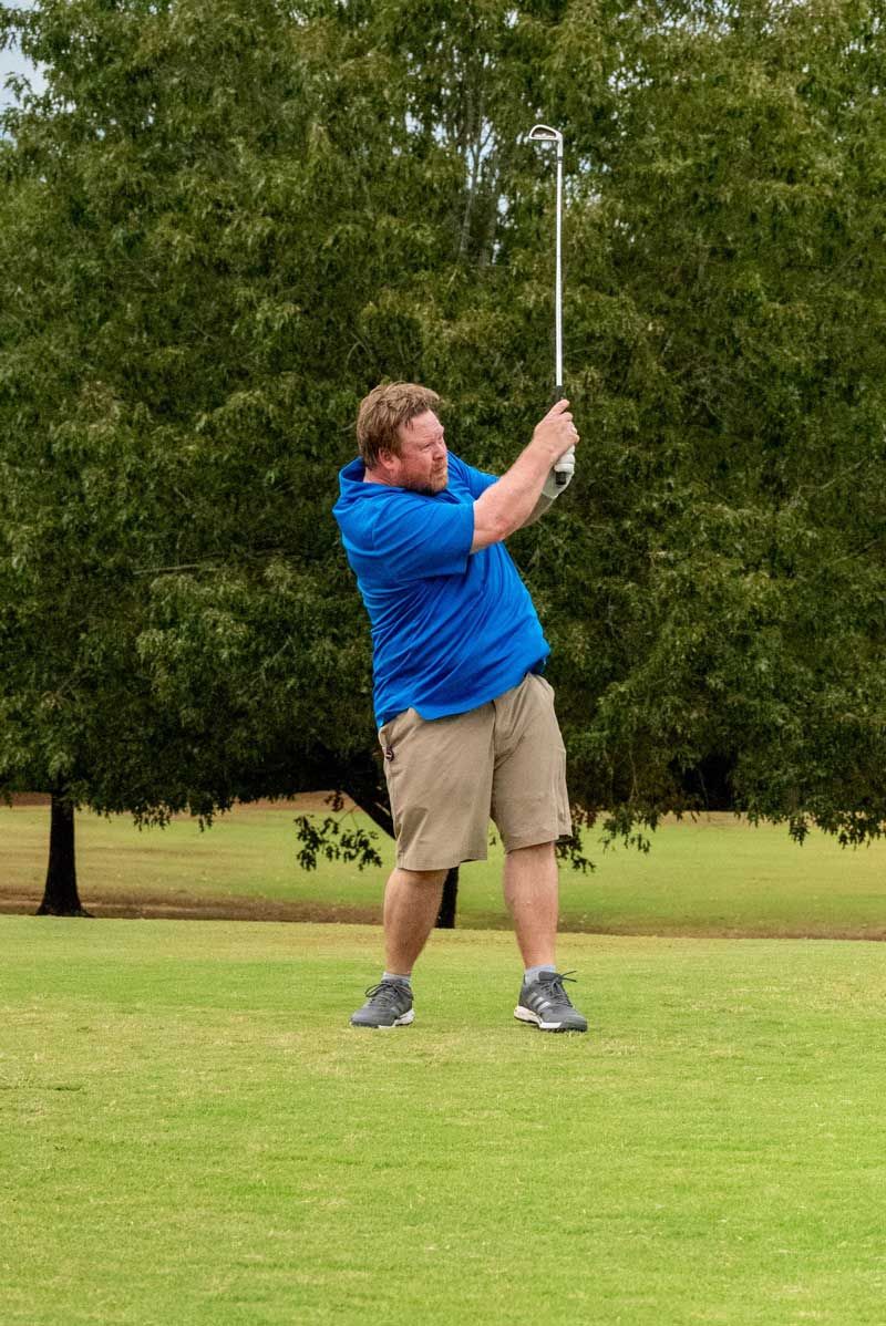 Man in blue shirt and khaki shorts swings a golf club on a green course.