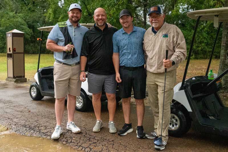Four men standing by golf carts on a rainy golf course, smiling.