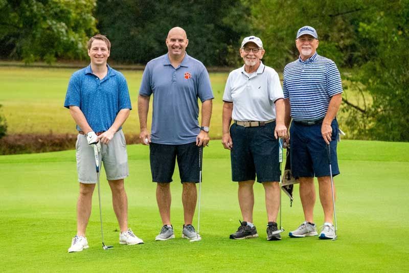 Four men pose on a golf course. They wear casual golf attire, holding clubs near a green with a flag.