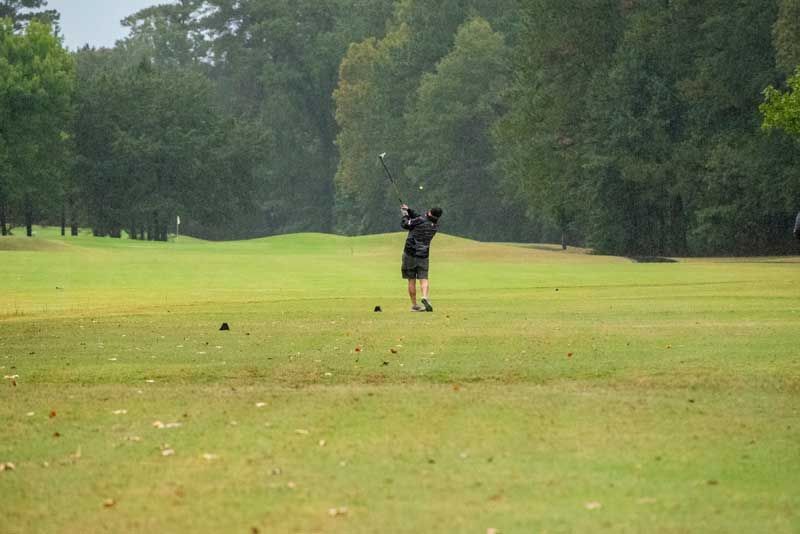 Golfer swings club on green course, trees in background, overcast weather.