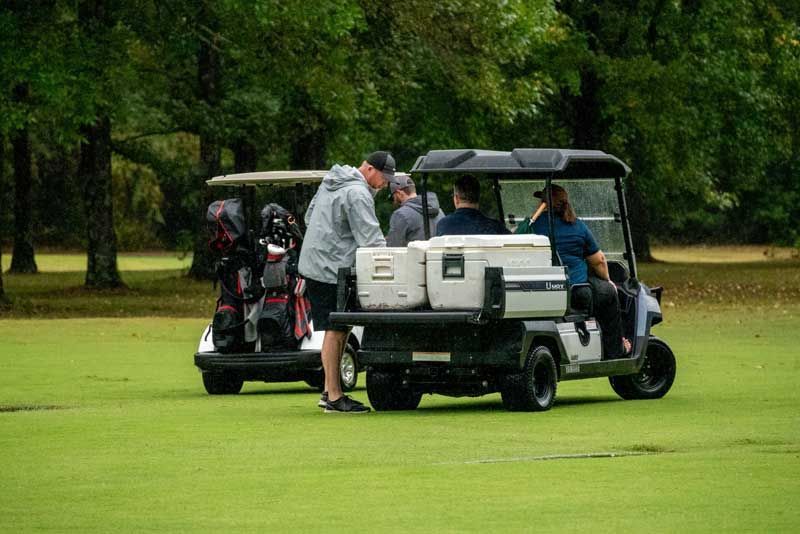 Two golf carts on a green golf course. People are loading a cooler into a cart.