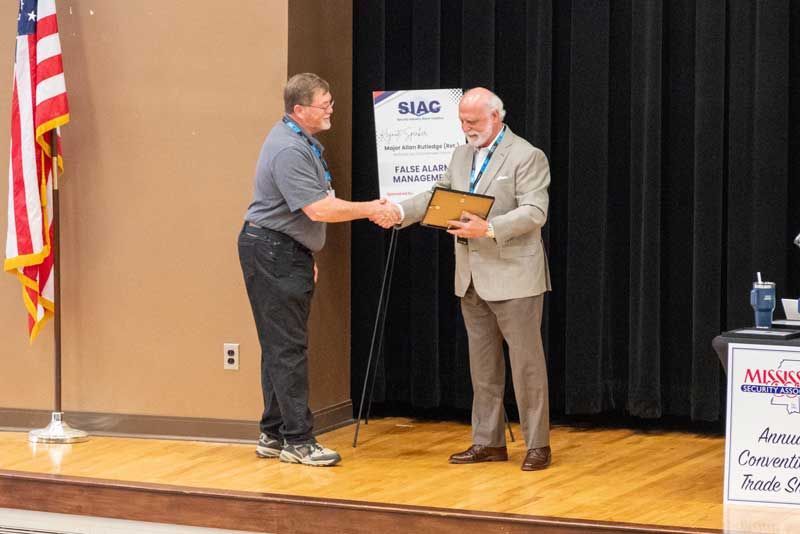Man receives an award at a conference. They shake hands on a stage. American flag and a sign in the background.