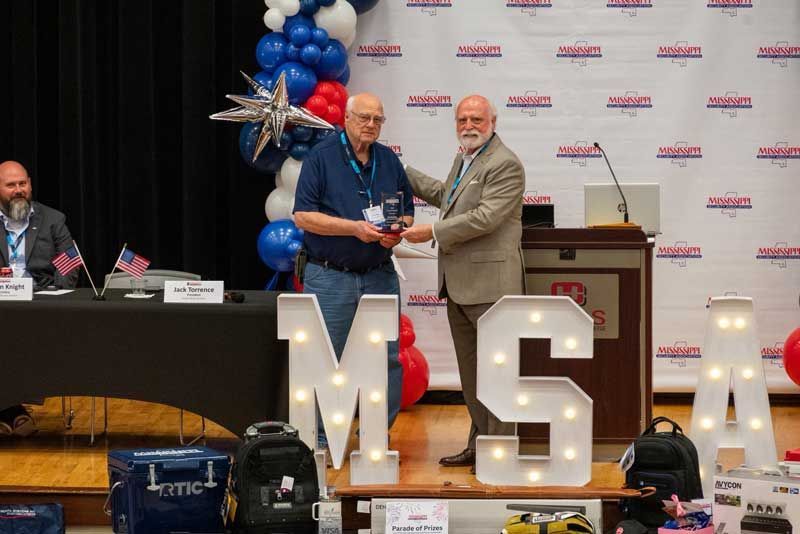 Man receiving award at event with MSA letters lit up. Balloons and table with flags are in the background.