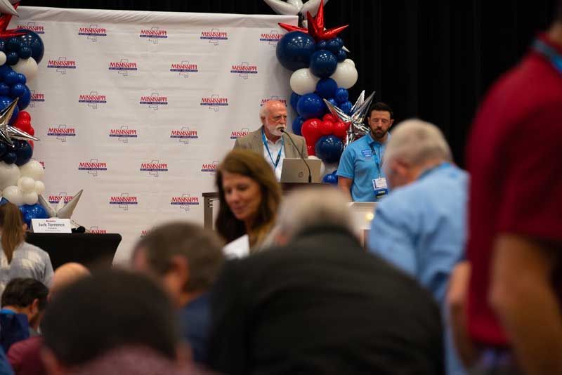 Man speaking at a podium in front of a backdrop with a crowd; balloons are visible.