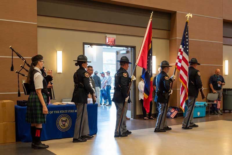 Honor guard presents flags at an event inside a building. A bagpiper plays nearby.