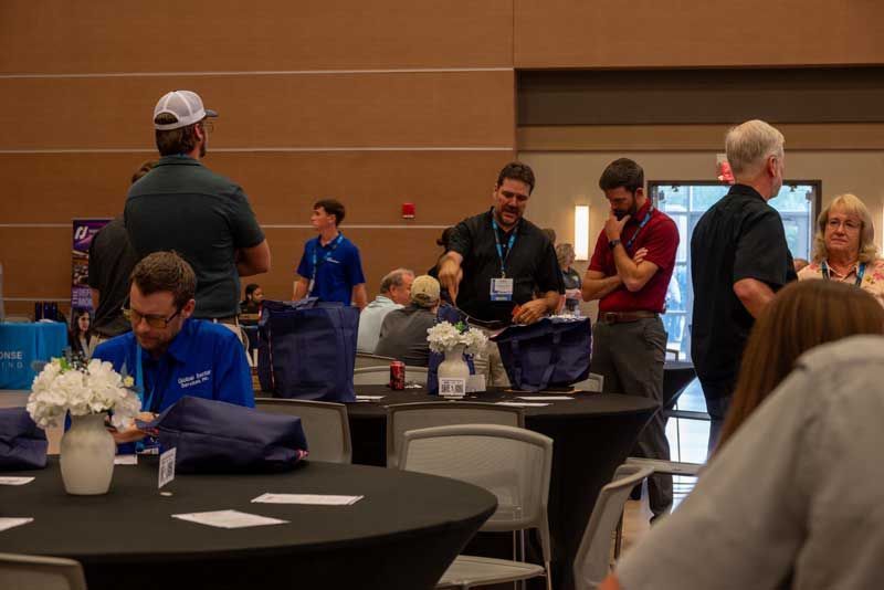 People at a conference. Several stand near tables, some look at items. Others sit.