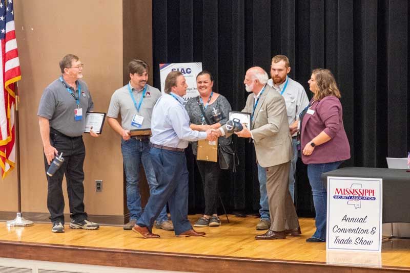 Group of people on stage shaking hands at an awards ceremony, holding plaques. American flag in background.
