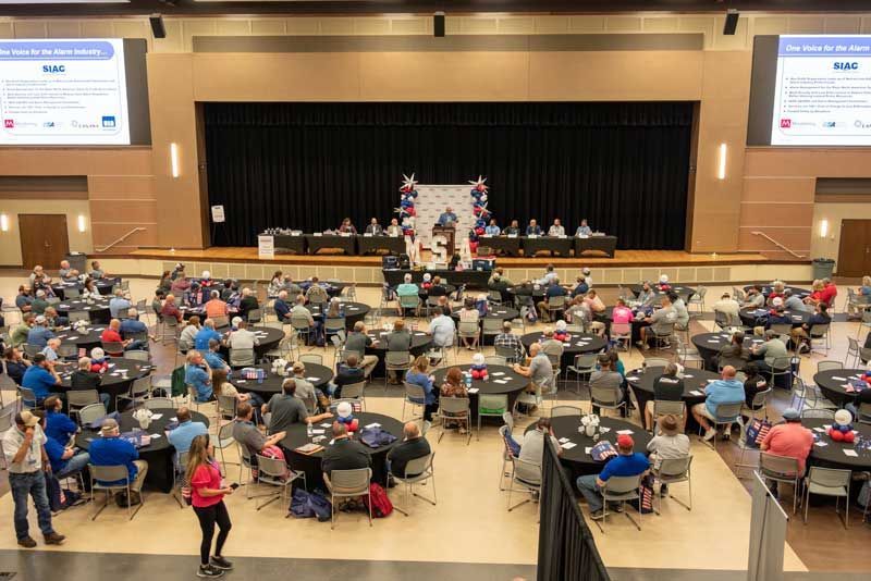 Large event with people seated at round tables. A stage has people at a podium and behind tables.