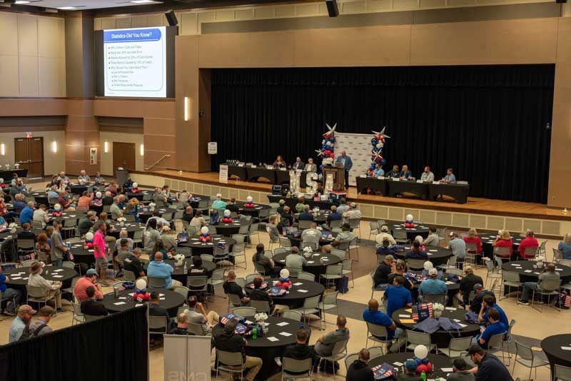 Large event in a hall; people seated at round tables facing a stage with speakers and a screen.